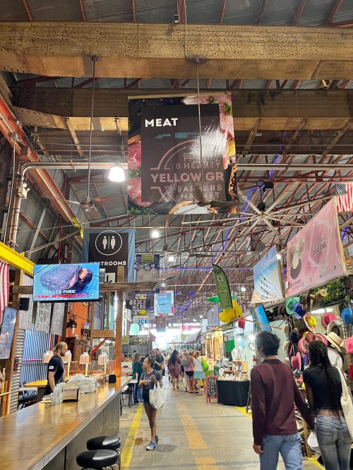 A bustling market interior with a "MEAT" sign, vendors, and people shopping in a vibrant atmosphere.