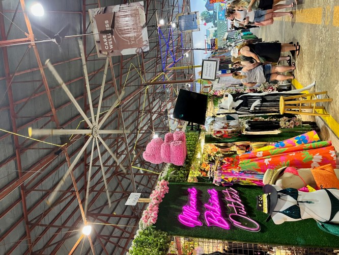 A vibrant market scene featuring colorful clothing stalls, a large fan, and a neon sign for "Miami Bikini Outlet."