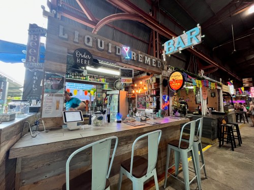 A vibrant bar named "Liquid Remedy" with colorful lights, drinks, and a rustic wooden counter in a lively setting.