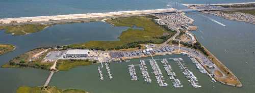 Aerial view of a marina with boats, surrounded by water and greenery, near a beach and a bridge in the background.