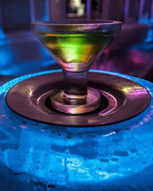 A colorful cocktail glass sits on a glowing blue surface, illuminated by vibrant lights.