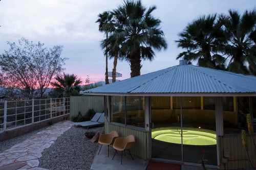 A modern outdoor space featuring a hot tub, palm trees, and a sunset sky in the background.