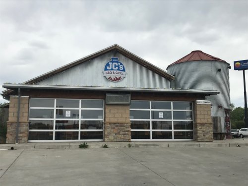 Exterior of JC's BBQ & Grill, featuring a stone facade, large windows, and a silos-style structure. "Open" sign visible.