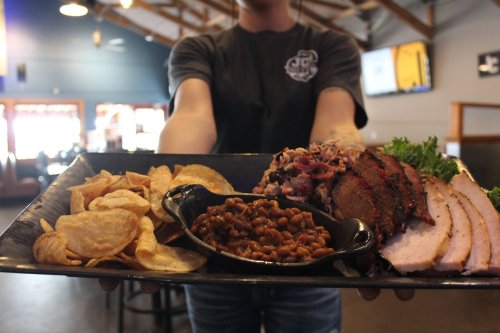 A person holds a platter with sliced meats, baked beans, and potato chips in a casual dining setting.