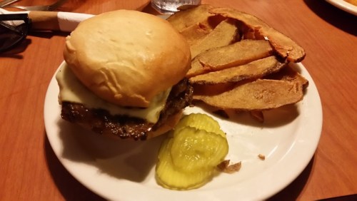 A plate with a cheeseburger, pickles, and crispy potato wedges on a wooden table.
