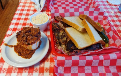 A plate of barbecue with sliced bread and pickles, alongside a bowl of baked beans on a checkered tablecloth.