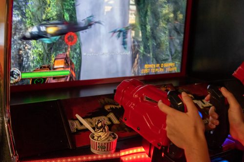 A person holds a red arcade gun while playing a shooting game, with a dessert cup nearby and a jungle scene on the screen.