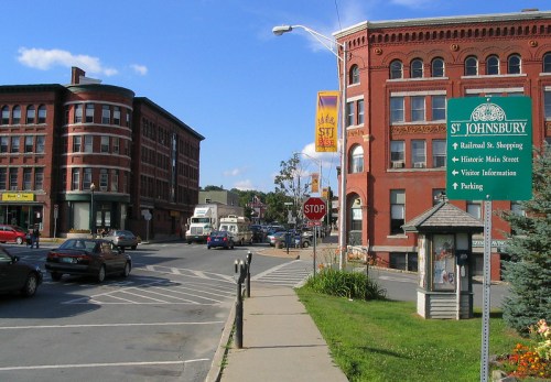 A street view of St. Johnsbury, featuring brick buildings, a stop sign, and directional signs for shopping and visitor info.