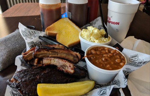 A plate of barbecue ribs, brisket, baked beans, potato salad, toast, and pickles, with drinks in the background.