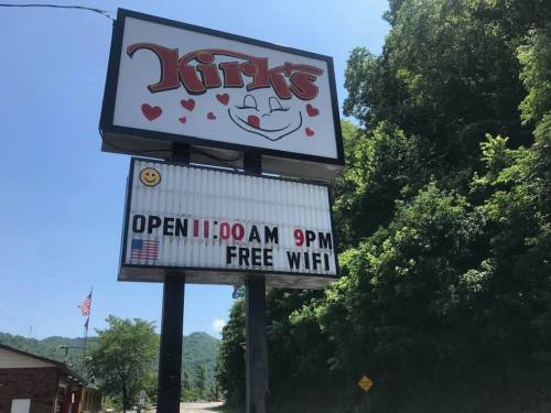 Sign for Kirk's with a smiling face, stating "Open 11:00 AM - 9:00 PM" and "Free WiFi," surrounded by greenery.