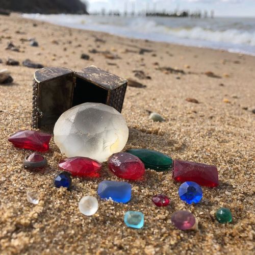 A collection of colorful gemstones and a glass orb scattered on sandy beach near a small, open box.