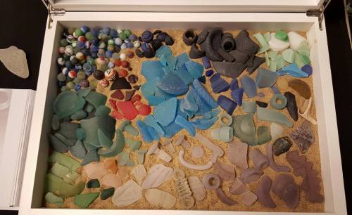 A collection of colorful sea glass and marbles arranged in a box filled with sand.