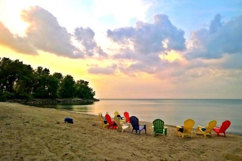 Colorful Adirondack chairs lined up on a sandy beach, facing a calm lake under a cloudy sunset sky.
