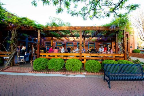 Outdoor dining area with people seated at wooden tables, surrounded by greenery and a brick walkway.