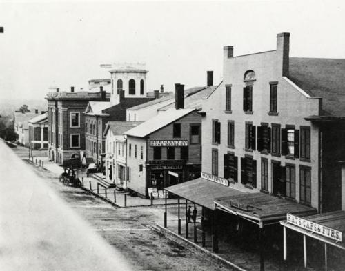 Historic black-and-white photo of a street lined with buildings, featuring shops and a tower in the background.