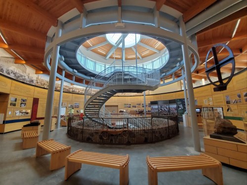 Interior of a museum featuring a spiral staircase, circular skylight, and exhibits along the walls. Wooden benches are visible.
