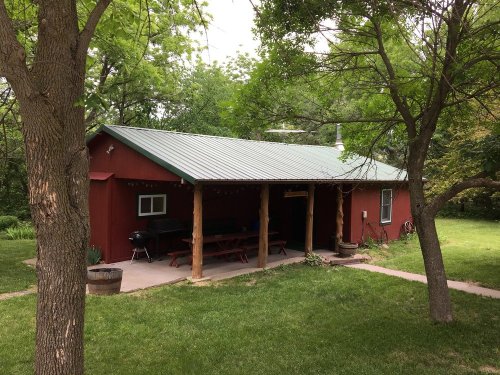 A red cabin with a green metal roof, surrounded by trees and grass, featuring a porch and picnic area.