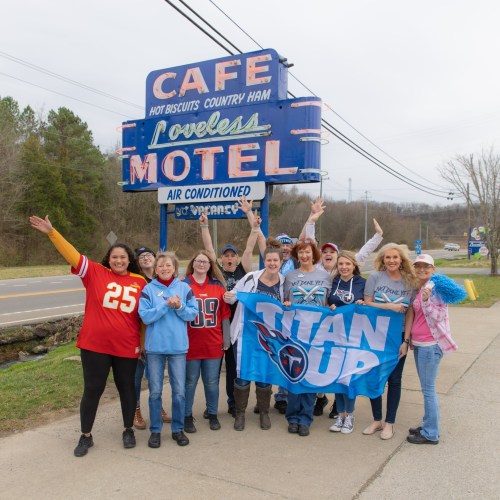 A group of enthusiastic fans in team jerseys poses in front of the Loveless Cafe and Motel sign, celebrating together.