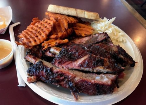 A plate of smoked ribs with sweet potato fries, coleslaw, and a slice of bread on a table.