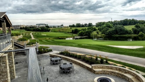 A scenic view of a golf course with lush green fairways, a pond, and cloudy skies, seen from a patio area.