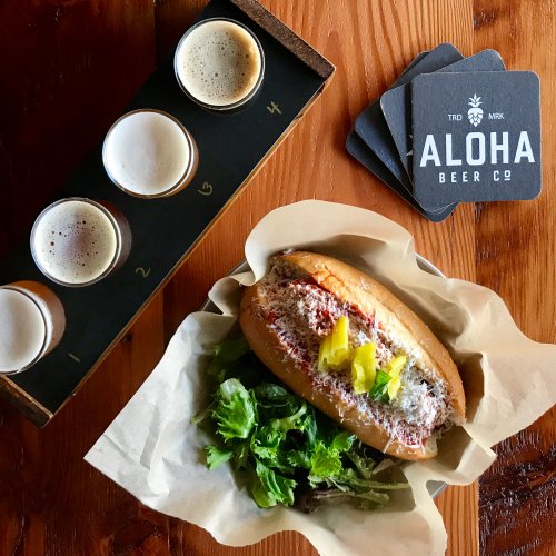 A hot dog topped with sauce and pickles, served with a side of greens, alongside a flight of beer samples.