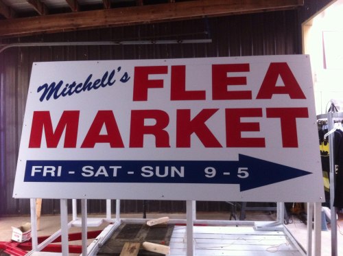 Sign for Mitchell's Flea Market, open Friday to Sunday from 9 AM to 5 PM, with an arrow pointing to the right.