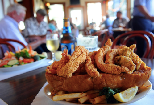 A plate of fried seafood on a bun with fries, lemon, and a beer, set in a busy restaurant with diners in the background.
