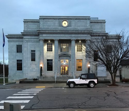 Historic stone building with a clock, flanked by trees, and a parked white Jeep in front, under a cloudy sky.