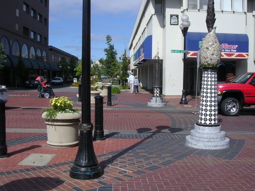 A vibrant urban street scene with decorative pavement, planters, and people walking, featuring unique sculptures and a red truck.