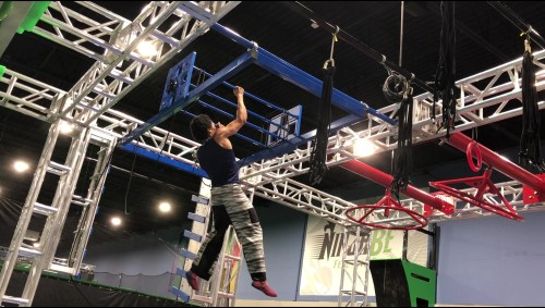 A person hangs from a blue structure in an indoor obstacle course, showcasing athleticism and strength.
