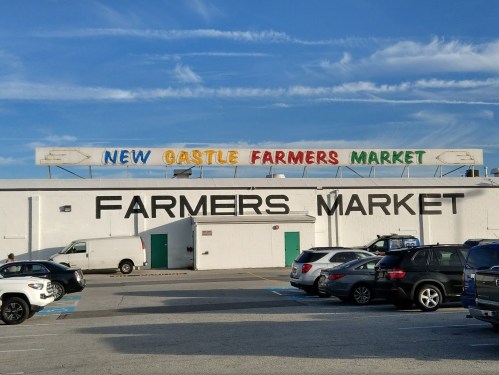 Sign for New Castle Farmers Market on a building, with parked cars in the foreground and a blue sky above.