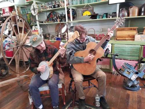 Two musicians play instruments in a cozy, vintage shop filled with antiques. One plays a banjo, the other an acoustic guitar.