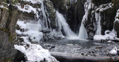 A frozen waterfall surrounded by snow-covered rocks and icy formations, with a calm river flowing below.