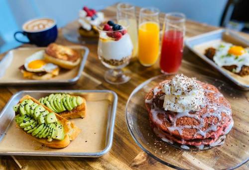 A vibrant brunch spread featuring waffles, avocado toast, parfait, and colorful drinks on a wooden table.