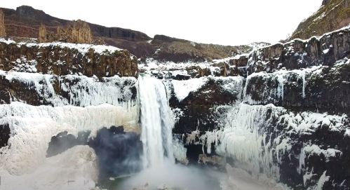A stunning waterfall surrounded by icy cliffs and snow, cascading into a misty pool below.
