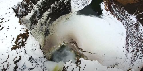 Aerial view of a frozen waterfall surrounded by snow and rocky terrain, with a partially frozen pool below.