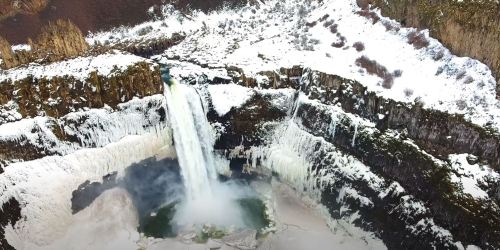 Aerial view of a frozen waterfall surrounded by snow-covered cliffs and icy terrain.