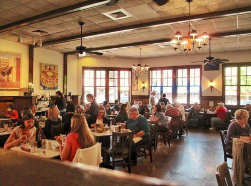 A bustling restaurant interior with diners at tables, warm lighting, and large windows creating a cozy atmosphere.