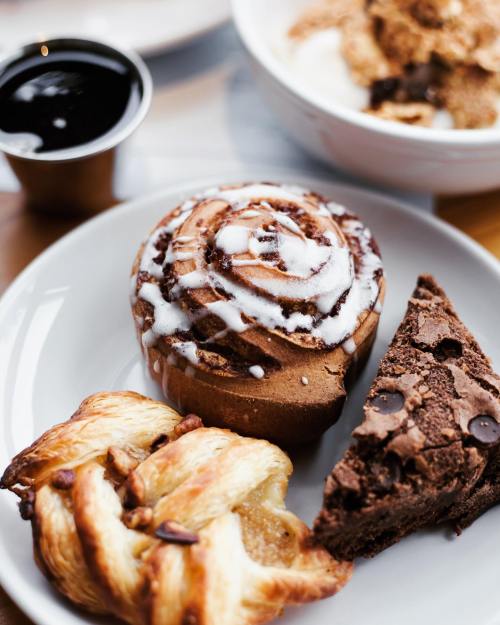 A plate with a cinnamon roll, a chocolate brownie, and a flaky pastry, accompanied by a small cup of syrup.