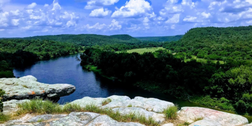A scenic view of a river winding through lush green hills under a bright blue sky with fluffy clouds.