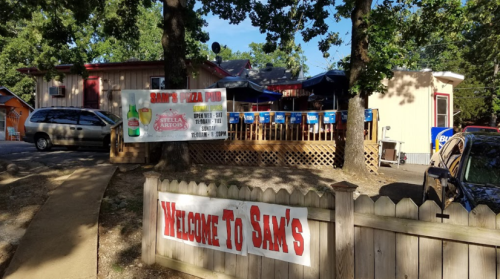 A welcoming sign for Sam's Pizza Pub, featuring outdoor seating and a menu board, surrounded by trees.