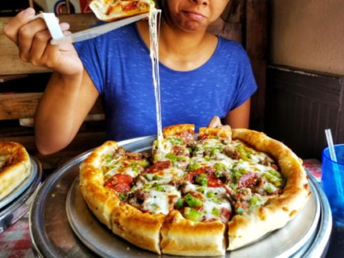 A person holds a slice of cheesy pizza, with toppings and a stuffed crust, looking pleased at a table.