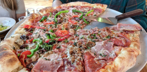 A large pizza topped with various meats, vegetables, and herbs, served on a round platter.