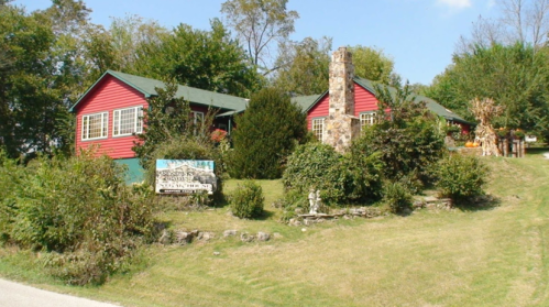 A red house with a stone chimney surrounded by greenery and a sign in front, set on a grassy hillside.