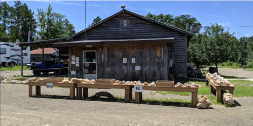 A rustic wooden building with tables displaying rocks for sale, set in a rural area with trees and vehicles nearby.