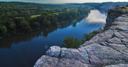 A serene river flows through lush green hills, viewed from a rocky cliff edge under a clear sky.