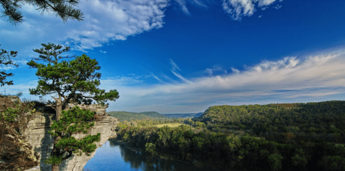A scenic view of a river winding through lush green hills under a blue sky with scattered clouds.
