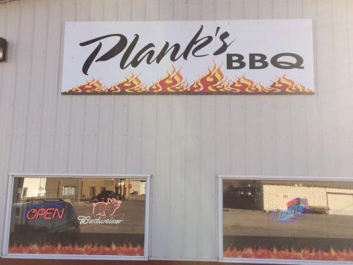 Sign for Plank's BBQ featuring flames, with "OPEN" and Budweiser logos in the windows.