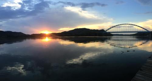 Sunset over a calm river, reflecting clouds and a bridge in the background, with hills silhouetted against the sky.
