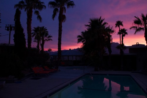 A serene poolside scene at dusk, with palm trees silhouetted against a vibrant purple and orange sky.